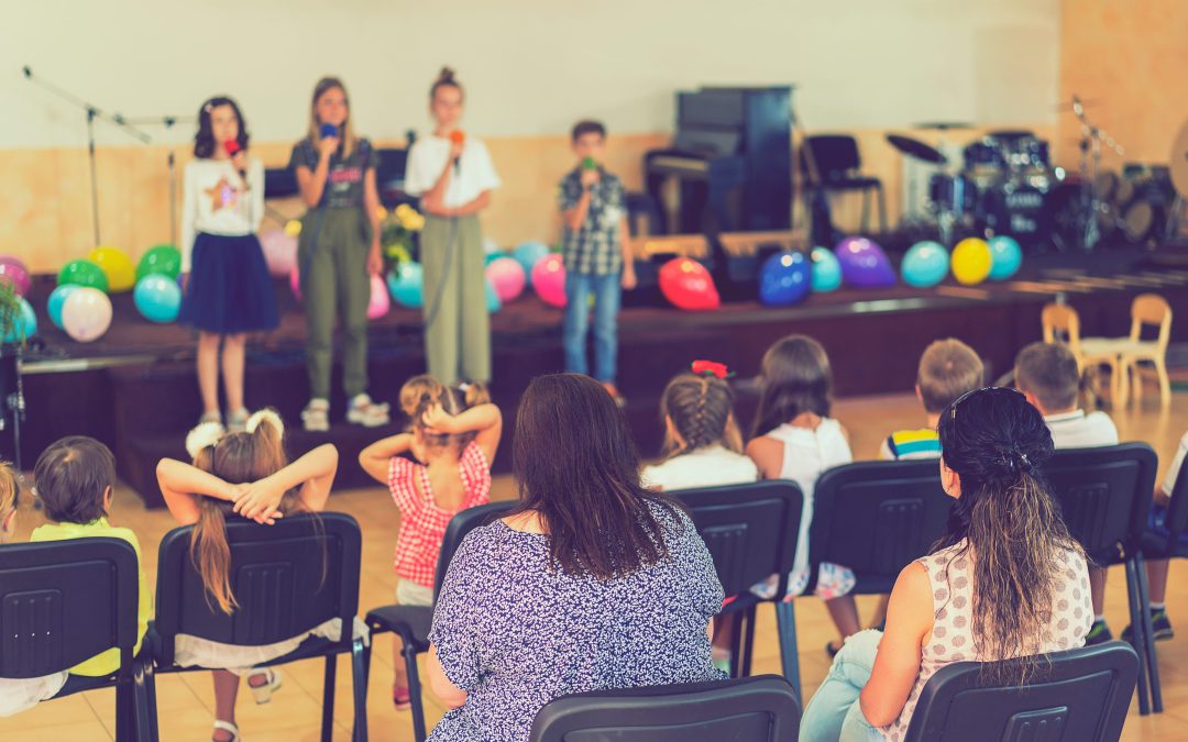 Young vocalists performing on stage with microphones in front of an audience of parents and children sitting in chairs, referencing